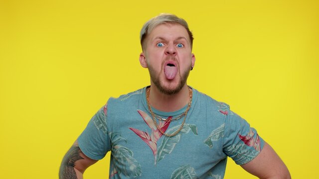 Attractive funny portrait of bearded adult guy in t-shirt showing tongue making faces at camera, fooling around, joking, aping with silly face, teasing. Young tourist man on yellow studio background