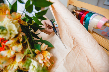 A florist cuts packaging paper with scissors on a desktop in a flower shop. On the paper is a bouquet of fresh flowers