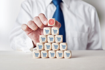 Businessman placing cube with target symbol on top of tiered wooden cubes with shopping cart icon.