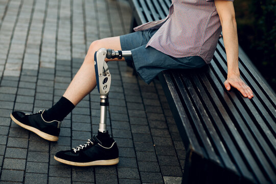 Disabled young man with leg prosthesis sitting on bench outdoor