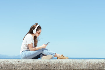 Young woman wearing headphones and checking her phone at the coastline in a sunny summer day.