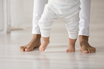 Crop close up of caring African American mom hold small toddler daughter hands make first steps...