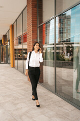 Woman with business backpack walking while speaking on the phone near a business building
