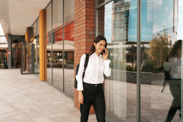 Woman with business backpack walking while speaking on the phone near a business building