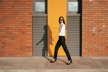 Young woman on high hills shoes and business wear walking confident along a brick building wall on a sunny day in sunglasses