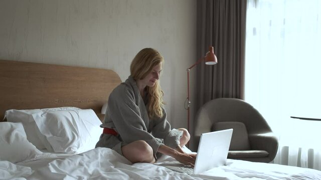 Caucasian woman in bathrobe working at laptop in hotel bed