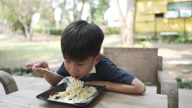Asian boy about 5 years wearing face mask under the chin eating spaghetti with boring expression at the restaurant
