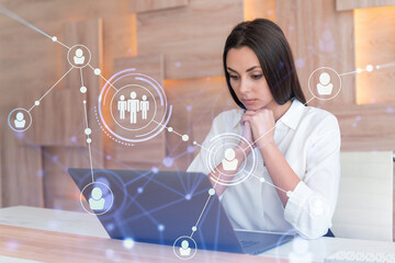 Attractive businesswoman in white shirt at workplace working with laptop to hire new employees for international business consulting. HR, social media hologram icons over office background