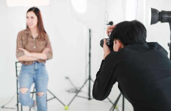 Selective focus on asian male photographer holding camera and taking photo of blur background long hair woman in studio with lighting equipments, tripods and white cutout.