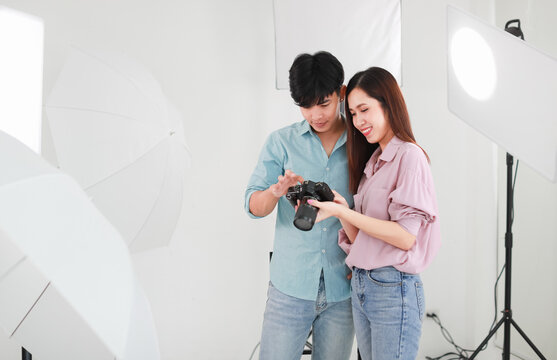 Portrait of two handsome man and beautiful woman wearing casual shirts, jeans, checking photos in camera while standing in indoor photo studio with white background cutout and lighting equipments.