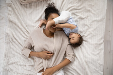 Top view of smiling young African American mother relaxing in bed at home with cute small baby daughter. Happy ethnic mom have fun rest in bedroom with little toddler kid child. Motherhood concept.