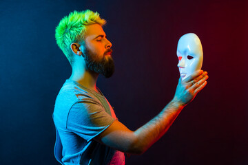 Side view of confident serious man in denim overalls, holding and looking at white mask in hands with attentive look, trying to understand hiding personality. Colorful neon light, indoor studio shot. - obrazy, fototapety, plakaty