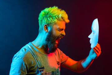 Profile of young adult attractive hipster man screaming angrily, holding white mask in hands,...