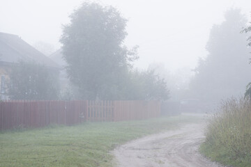 View of a deserted village street. Morning fog. Summer rural foggy landscape. Dirt road, fences and wooden houses. Everyday life in the countryside.