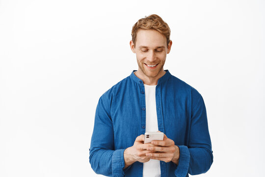 Handsome modern guy with red hair messaging, reading screen on smartphone, chatting and smiling, making online shopping order on phone, standing over white background