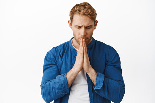 Hopeful serious redhead man, praying, holding hands in pray begging god, close eyes and suplicating, pleading about something, standing over white background