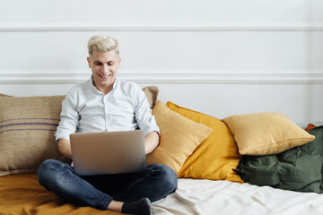 Young attractive smiling blonde guy is browsing at his laptop, sitting at home on the cozy sofa at home, wearing casual outfit