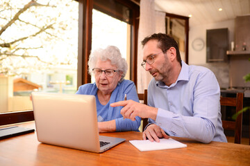 mature man helping elderly senior woman at home with paperwork and computer internet lesson - obrazy, fototapety, plakaty