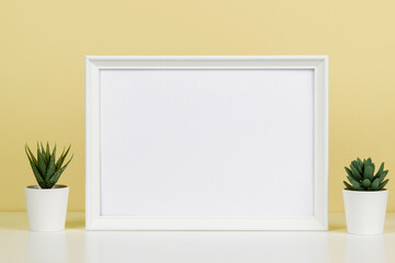 White blank wooden mock up frame and small succulent flowers on white desk with light yellow background. Front view