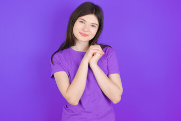 Charming serious young beautiful Caucasian girl wearing purple T-shirt over purple background  keeps hands near face smiles tenderly at camera