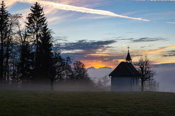 Fototapeta premium Kapelle im Winter bei Sonnenuntergang, im Hintergrund das Rheintal mit Nebelmeer und der Säntis im orangen Sonnenlicht. chapel in sunset with sea of fog and colored sky
