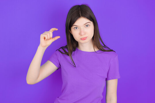 young beautiful Caucasian girl wearing purple T-shirt over purple background purses lip and gestures with hand, shows something very little.