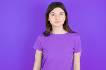 Fototapeta premium Stunned young beautiful Caucasian girl wearing purple T-shirt over purple background stares reacts on shocking news. Astonished girl holds breath