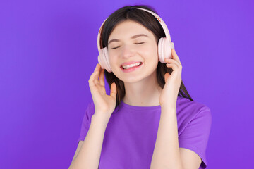 young beautiful Caucasian girl wearing purple T-shirt over purple background with headphones on her head, listens to music, enjoying favourite song with closed eyes, holding hands on headset.