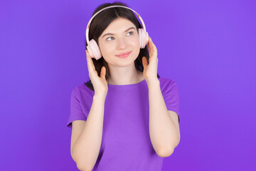 Fototapeta premium young beautiful Caucasian girl wearing purple T-shirt over purple background wears stereo headphones listens music concentrated aside. People hobby lifestyle concept