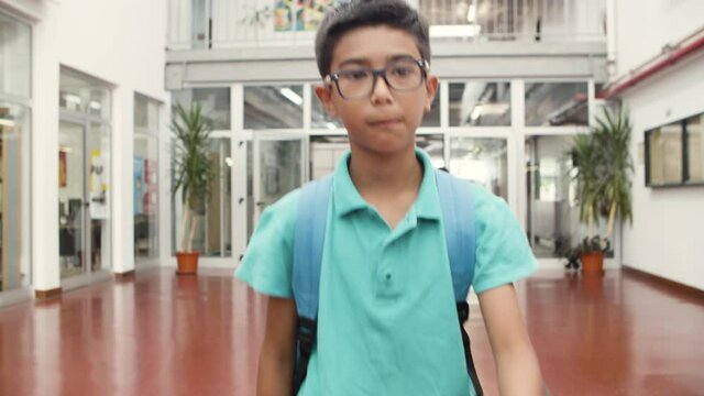 Medium shot of smiling boy in eyeglasses walking in school. Excited schoolboy going to class after vacation, looking around and at camera. Schoolgirls going behind and talking. School time concept.