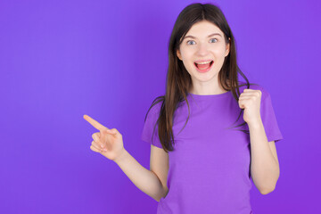 young beautiful Caucasian girl wearing purple T-shirt over purple background points at empty space holding fist up, winner gesture.