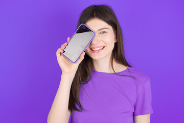 Fototapeta premium young beautiful Caucasian girl wearing purple T-shirt over purple background holding modern smartphone covering one eye while smiling