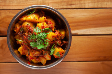 Close-up of Indian vegetarian dish of spicy Potato and Tomato curry garnished with green coriander fresh leaves. Served in a steel bowl over wooden background.