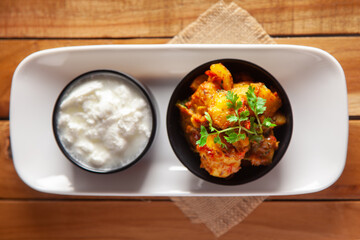 Close-up of Indian vegetarian dish of spicy Potato and Tomato curry garnished with green coriander fresh leaves with curd. over wooden background.
