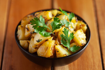 Close-up of Indian vegetarian classic dish Jeera Aloo - Potatoes Flavored with Cumin  garnished with green coriander fresh leaves. Served in black ceramic bowl over wooden background