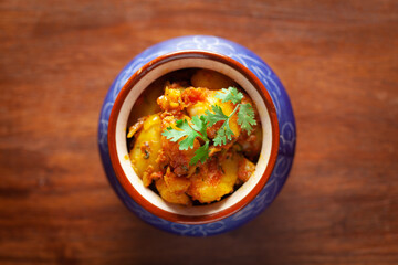 Close-up of Indian vegetarian dish of spicy Potato and Tomato curry garnished with green coriander fresh leaves. Served in a ceramic handi (clay pot) over wooden background.