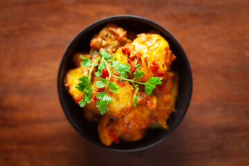 Close-up of Indian vegetarian dish of spicy Potato and Tomato curry garnished with green coriander fresh leaves. Served in a black ceramic bowl over wooden background.