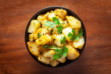 Close-up of Indian vegetarian classic dish Jeera Aloo - Potatoes Flavored with Cumin  garnished with green coriander fresh leaves. Served in black ceramic bowl over wooden background