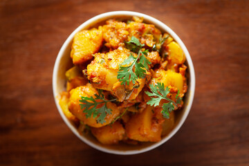 Close-up of Indian vegetarian dish of spicy Potato and Tomato curry garnished with green coriander fresh leaves. Served in a white ceramic bowl over wooden background.