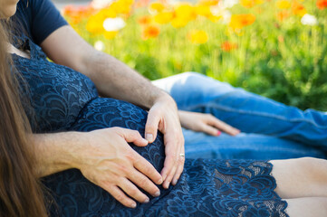 Eine schwangere Frau sitzt in einer Blumenwiese und wird von ihrem Partner liebevoll umarmt. Er...