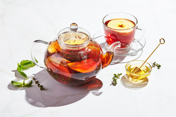 Fruit tea with apples and thyme and honey in glass teapot and cup on white background with hard shadows