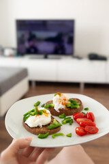 Close up of female hand holding a white plate with whole grain toasted bread and pouched eggs with cherry tomatoes and green peppers, interiour of living room, lifestyle concept