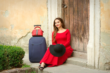 A young caucasian woman is sitting on the threshold of an old house in a red long dress with...