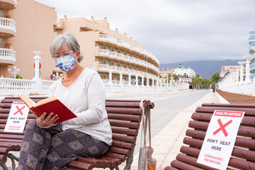 Fototapeta na wymiar Coronavirus. Senior woman sitting outdoor in a bench relaxes reading a book. Health notices about prevention of coronavirus