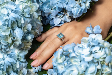 Fashion silver or white gold ring with a blue rectangular aquamarine stone on hand. Elegant topaz ring for women. Jewelry ring with a precious stone. Blue flowers background. - obrazy, fototapety, plakaty