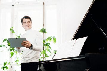 A confident person is a musician sitting near the piano. The gray-haired blogger is a positive man in age, writes down new cases on the project in the gadget. Portrait of a creative person smiling.