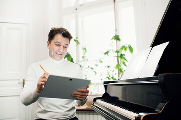 A male musician writes down the notes in a tablet for a new song. The artist works at home and plays the piano. He teaches at a music school. Professional activity of an adult.