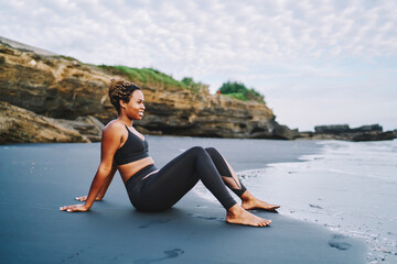 Fototapeta na wymiar Side view of sportive girl with casual figure resting at beach enjoying morning physical recreation, carefree dark skinned jogger in tracksuit resting at black sand and dreaming about body positive