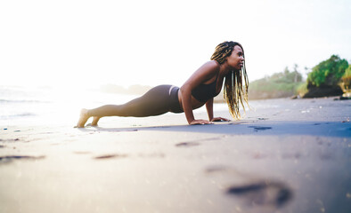 African American woman in sportive tracksuit doing push up exercising during morning workout training at seashore beach, strong fit girl with casual figure training body strength at coastline