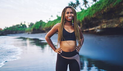 Obraz premium Cheerful African American sports woman with dreads standing at seashore beach and smiling, happy dark skinned woman dressed in tracksuit clothes for workout enjoying healthy lifestyle and vitality